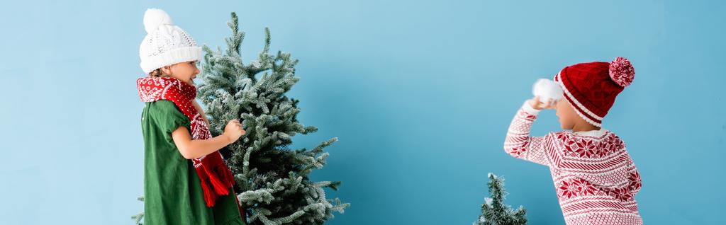 panoráma lövés a gyerekek téli ruhában játszik hógolyókat közelében karácsonyfák kék - Fotó, kép