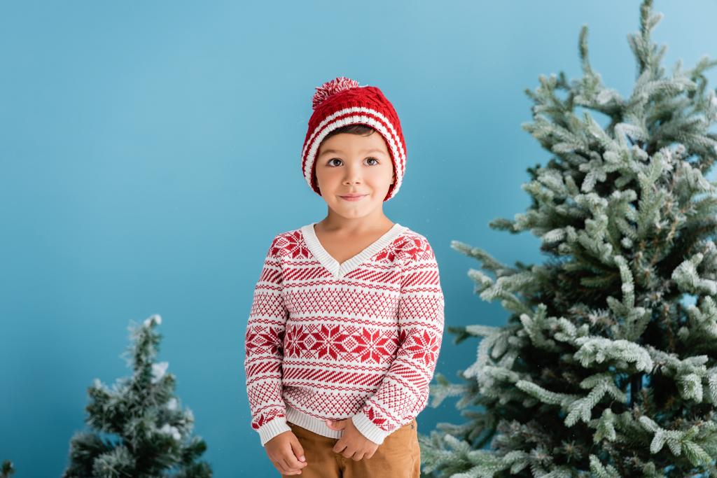 jongen in winter outfit staan in de buurt van kerstbomen op blauw - Foto, afbeelding