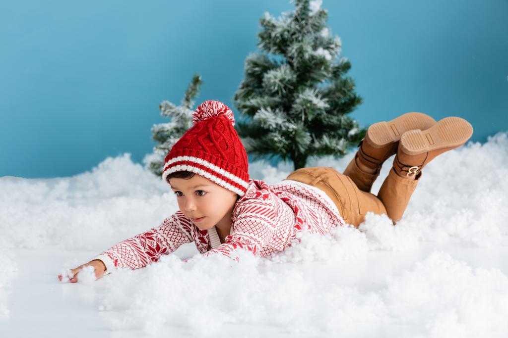 boy in winter outfit lying on white snow near christmas trees on blue - Photo, Image
