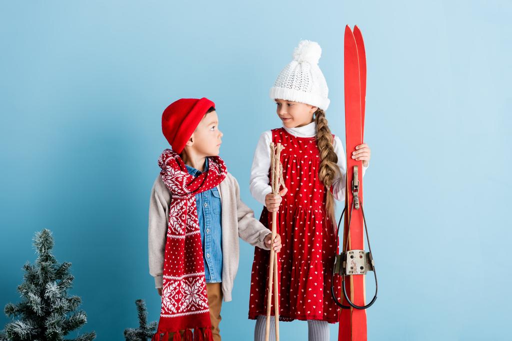 meisje in hoed kijken naar jongen terwijl het houden van skistokken en ski 's op blauw - Foto, afbeelding