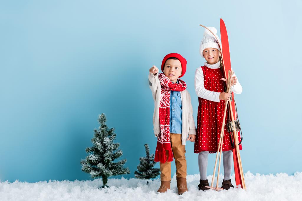 menino de chapéu e cachecol apontando com o dedo perto irmã segurando pólos de esqui e esquis, enquanto em pé sobre azul - Foto, Imagem