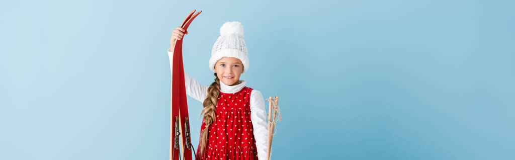 image horizontale d'enfant en tenue d'hiver debout avec bâtons de ski et skis isolés sur bleu - Photo, image