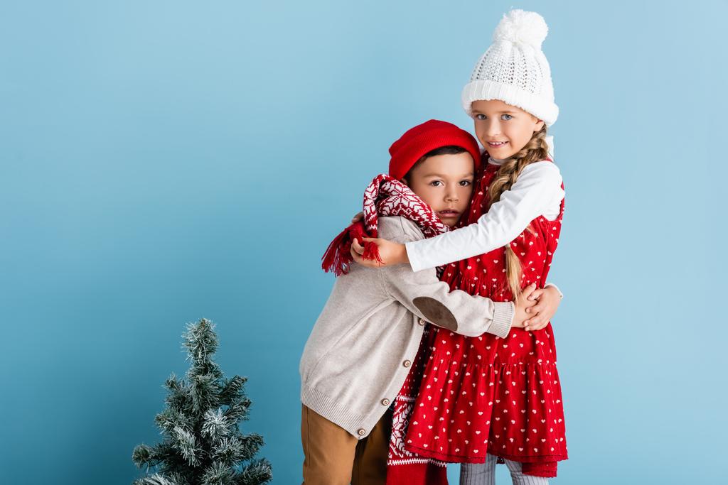 niños en traje de invierno abrazándose cerca del árbol de Navidad en azul - Foto, Imagen