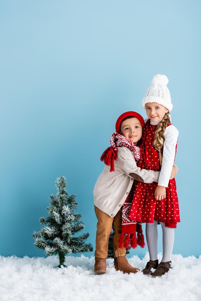Kinder im Winteroutfit umarmen sich in der Nähe des Weihnachtsbaums auf blauem Grund - Foto, Bild