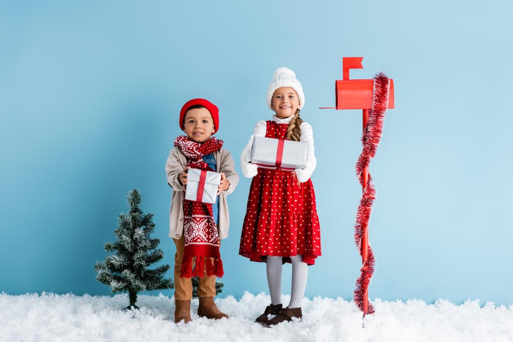 děti v zimním oblečení stojící na sněhu a držící dárky v blízkosti poštovní schránky a jedle na modré - Fotografie, Obrázek
