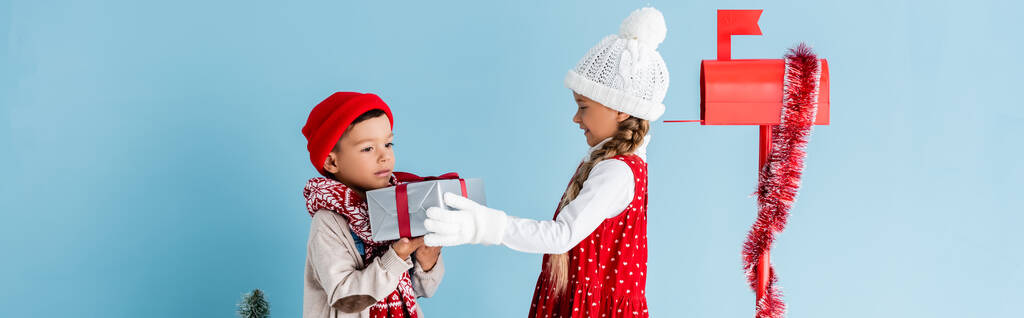 concepto panorámico de niño en traje de invierno celebración presente cerca de la hermana y buzón aislado en azul - Foto, imagen