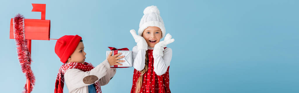 горизонтальний урожай хлопчика в зимовому вбранні, що тримає подарунок біля збудженої сестри і поштової скриньки ізольовані на синьому
 - Фото, зображення