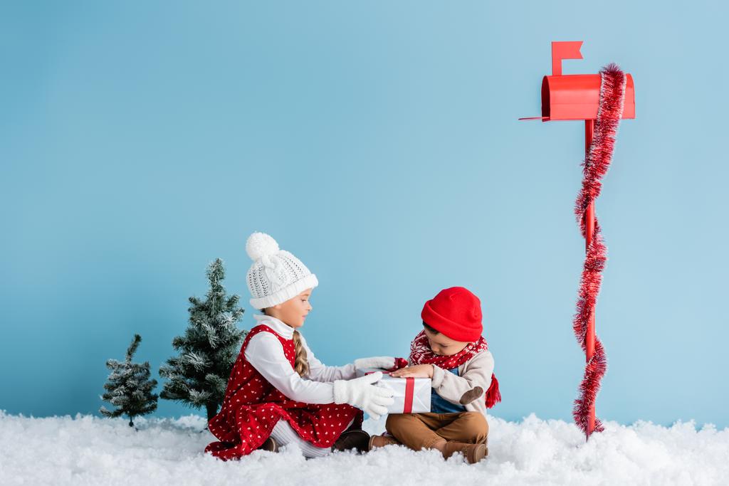 kids in winter outfit zitten op sneeuw en aanraken heden in de buurt brievenbus op blauw - Foto, afbeelding
