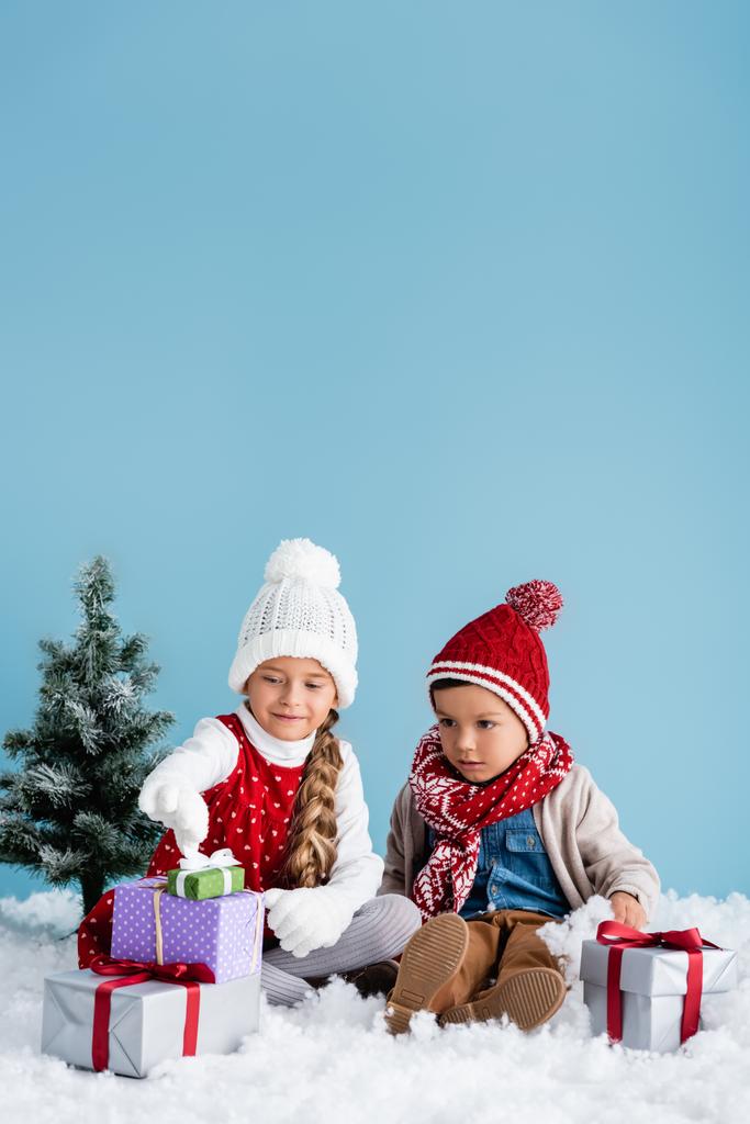 gyerekek téli ruhában ül a hóban, és nézi ajándékok közelében fenyő elszigetelt kék - Fotó, kép
