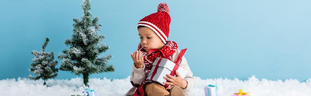panoramic concept of boy in hat and winter outfit sitting on snow and holding present while blowing on hand isolated on blue - Photo, Image
