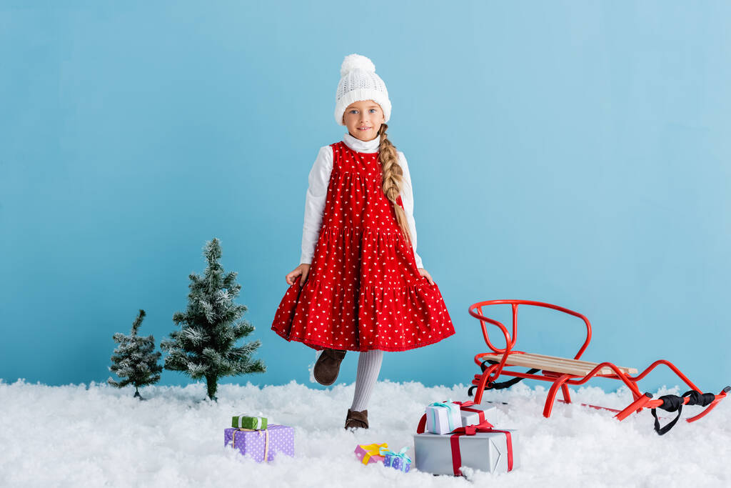 lapsi hattu ja talvi asu seisoo lumessa lähellä lahjoja ja reki eristetty sininen - Valokuva, kuva