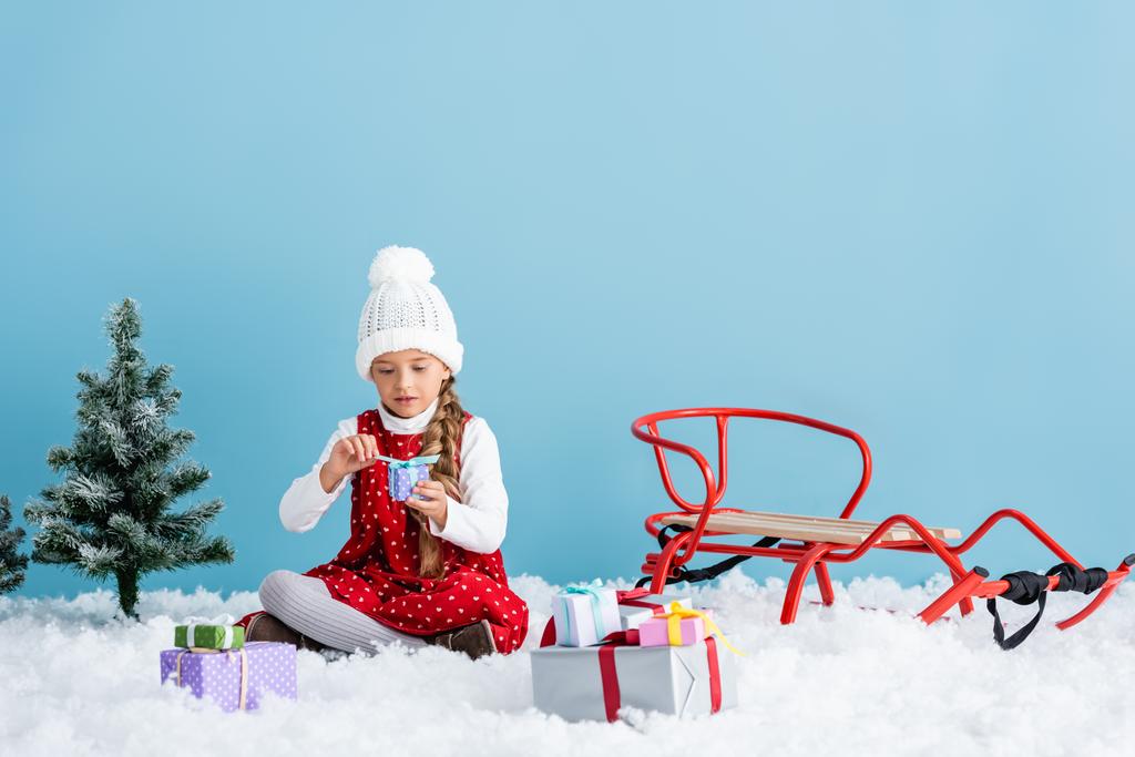 ребенок в шляпе и зимнем наряде сидит на снегу и держит подарок рядом сани изолированы на синий - Фото, изображение
