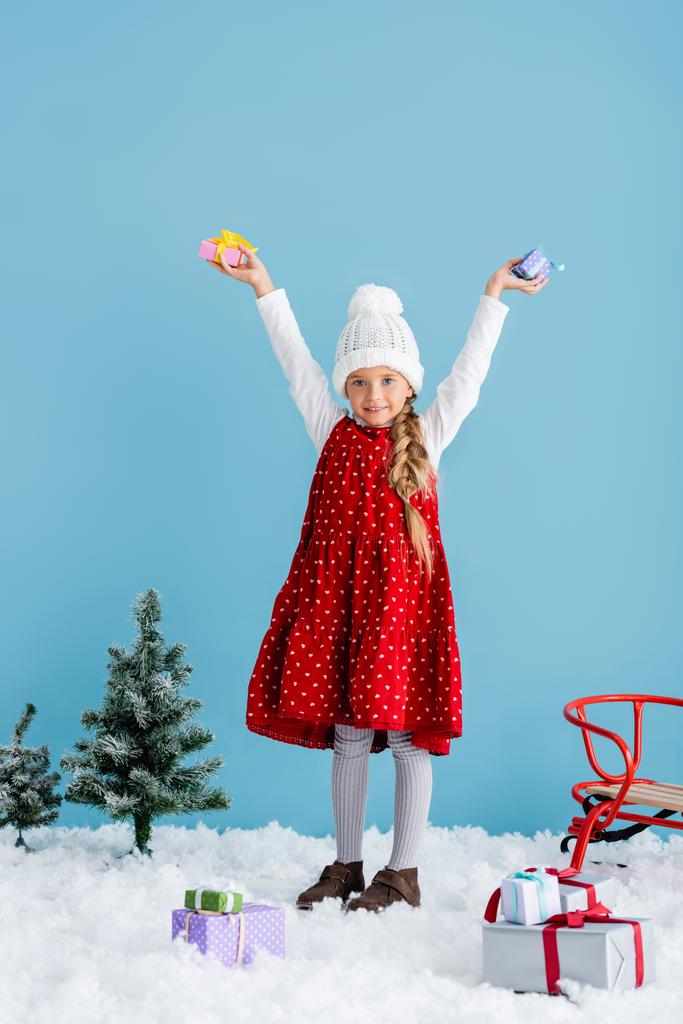 niño con sombrero y traje de invierno de pie sobre la nieve y la celebración de regalos por encima de la cabeza cerca del trineo aislado en azul - Foto, Imagen