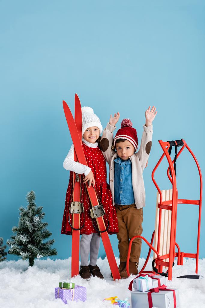 menina em roupas de inverno segurando esquis perto do irmão com as mãos acima da cabeça de pé perto de sleight e presentes na neve isolada em azul  - Foto, Imagem