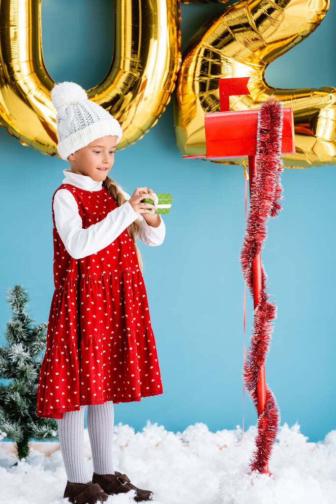 девушка в шляпе держит подарок, стоя возле почтового ящика и воздушных шаров на синий - Фото, изображение