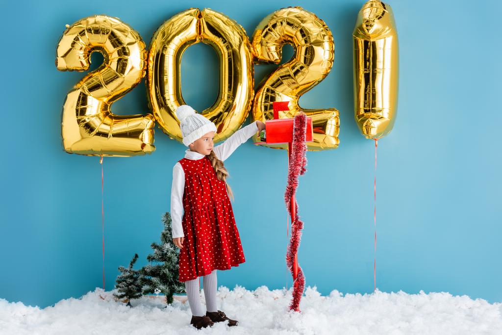 chica en sombrero tomando regalo de buzón cerca de globos con números, mientras que de pie en la nieve en azul - Foto, Imagen