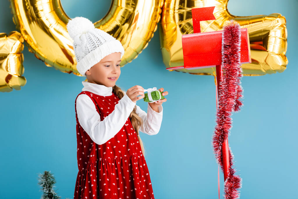 Şapkalı ve kışlıklı kız elinde kırmızı posta kutusunun yanındaki hediyeyi tutuyor.  - Fotoğraf, Görsel