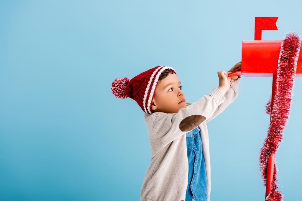 garçon en chapeau et tenue d'hiver atteignant boîte aux lettres rouge sur bleu  - Photo, image