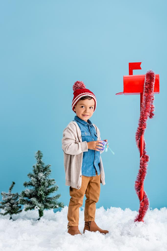 мальчик в зимнем наряде держит подарок возле красного почтового ящика, стоя на синем  - Фото, изображение