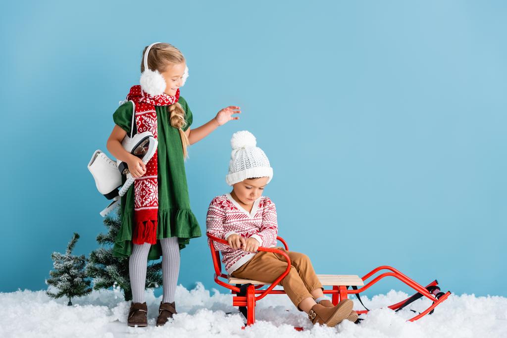 冬のイヤマフとスカーフの女の子はアイススケートで立っていると青にそりで男の子を見て - 写真・画像