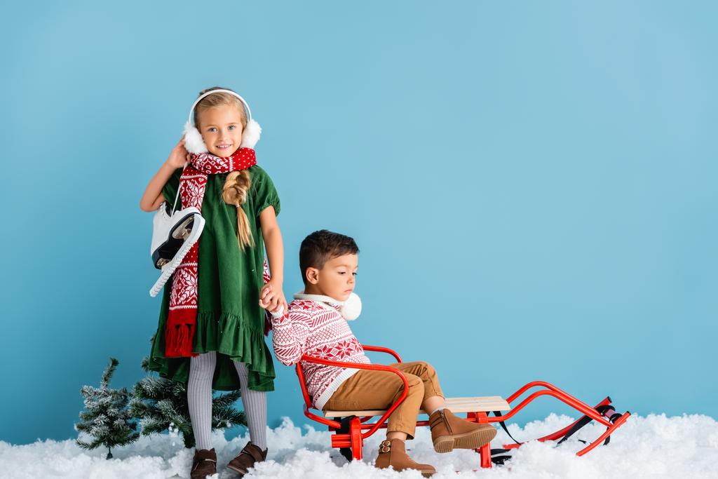 girl in winter earmuffs and scarf standing with ice skates and holding hands with boy in sleigh on blue - Photo, Image