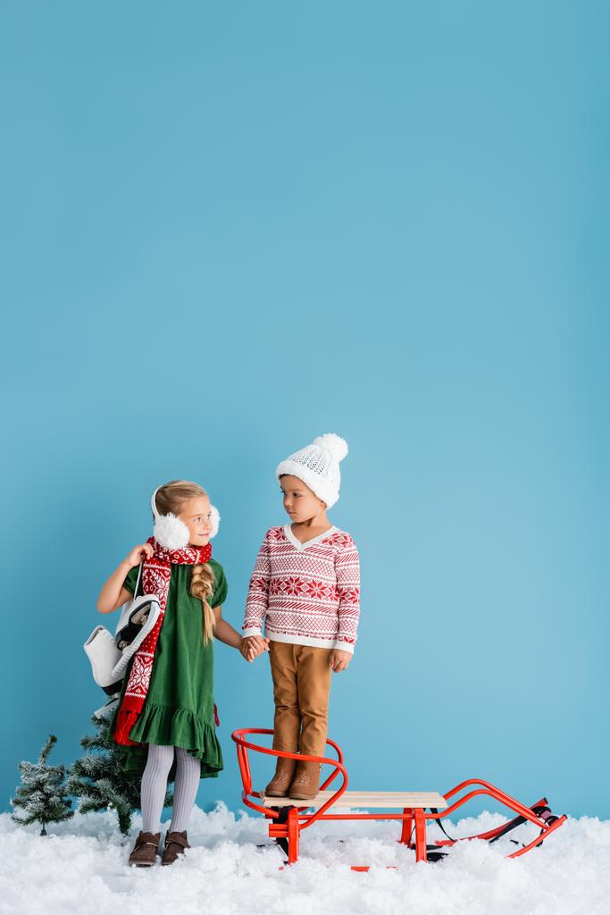 дівчина в зимових вухах і шарфі, що стоїть з льодовими ковзанами і тримає руки з хлопчиком в капелюсі біля саньйонів на синьому
 - Фото, зображення