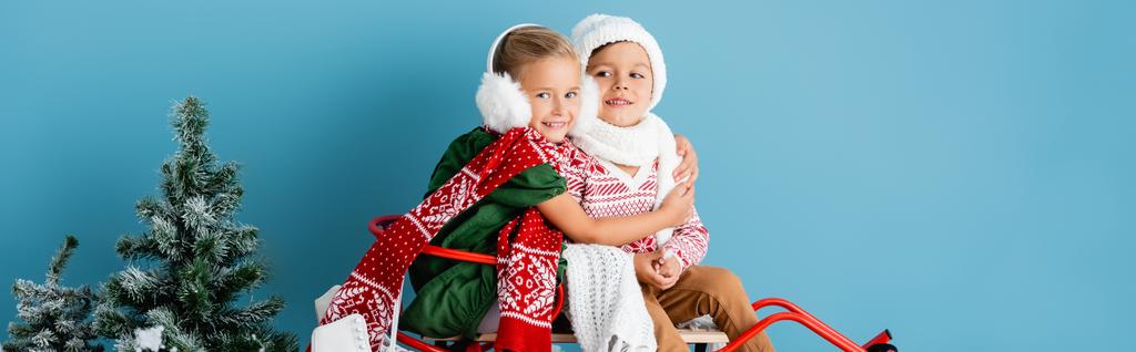 panoramic crop of girl in winter earmuffs sitting on sleigh and hugging brother in hat on blue - Photo, Image