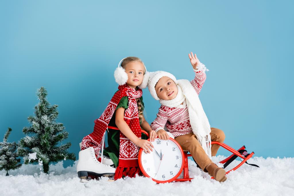 enfants en tenue d'hiver assis sur traîneau près de l'horloge, pins et patins à glace sur bleu - Photo, image
