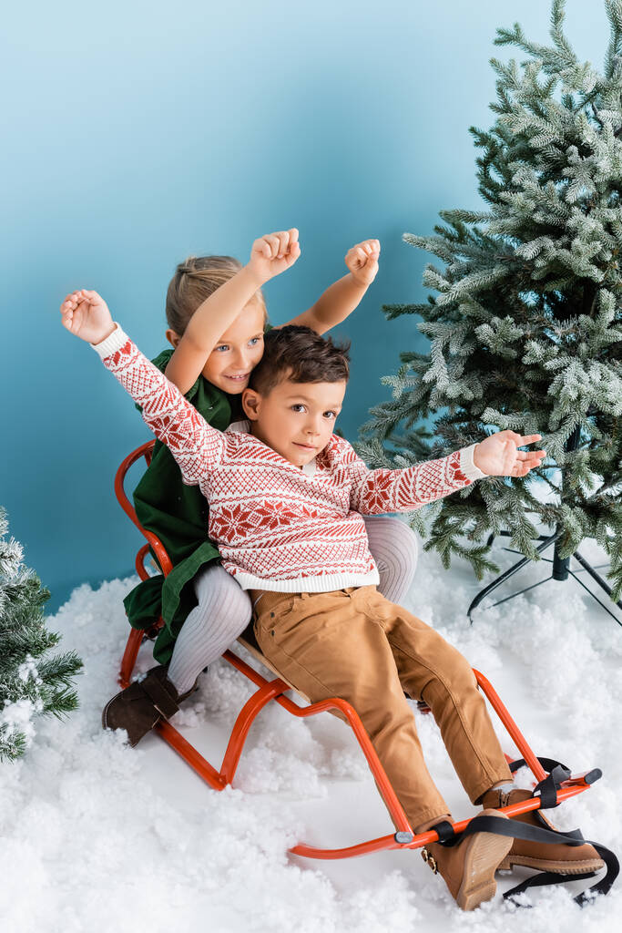 возбужденные дети с руками над головой сидят в санях на белом снегу рядом с зелеными соснами на голубом - Фото, изображение