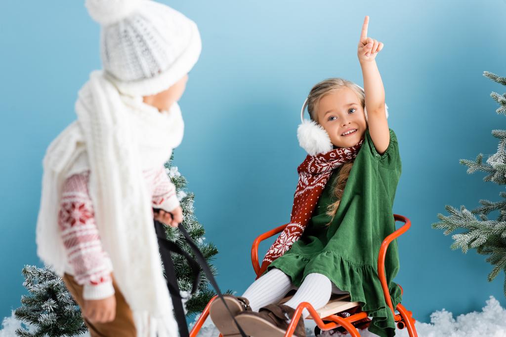 selective focus of girl pointing with finger while sitting on sleigh near boy on blue - Photo, Image