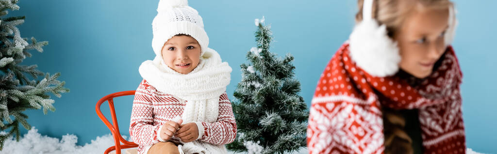 horizontaal gewas van jongen in winter hoed zitten op slee in de buurt zus op blauw - Foto, afbeelding
