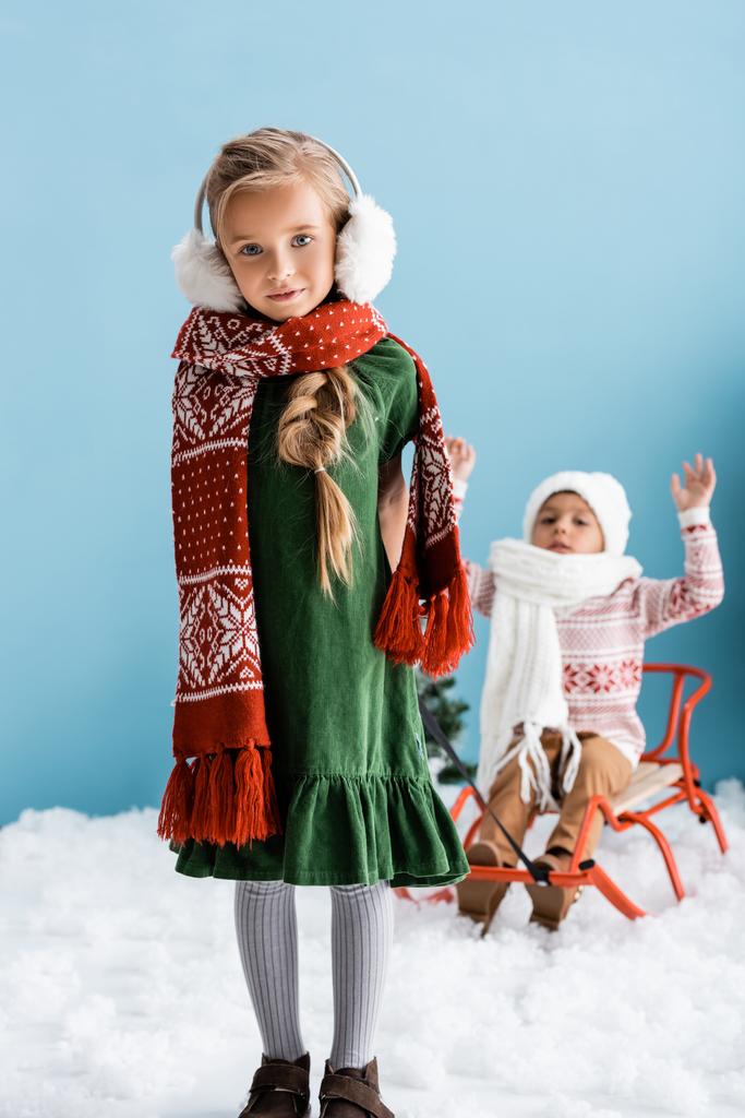 selektiver Fokus von Mädchen im Winteroutfit, die einem Jungen mit Hut auf einem Schlitten auf blauem Grund eine Fahrt geben - Foto, Bild