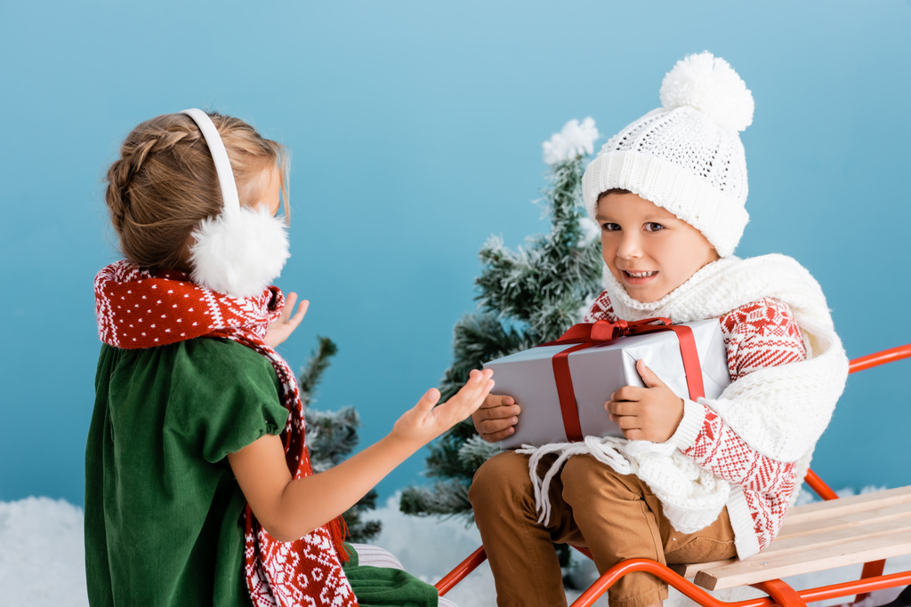 boy in knitted hat holding present while sitting on sleight near girl in winter earmuffs gesturing on blue - Photo, Image
