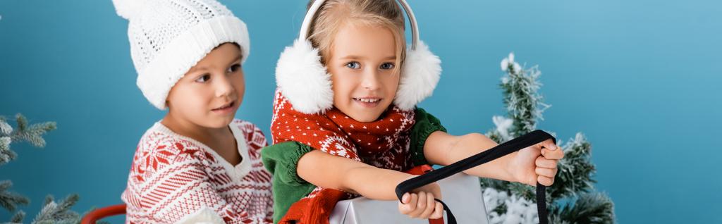 青い松の近くにプレゼントと一緒にそりに乗る冬の衣装の子供たちのパノラマ作物 - 写真・画像