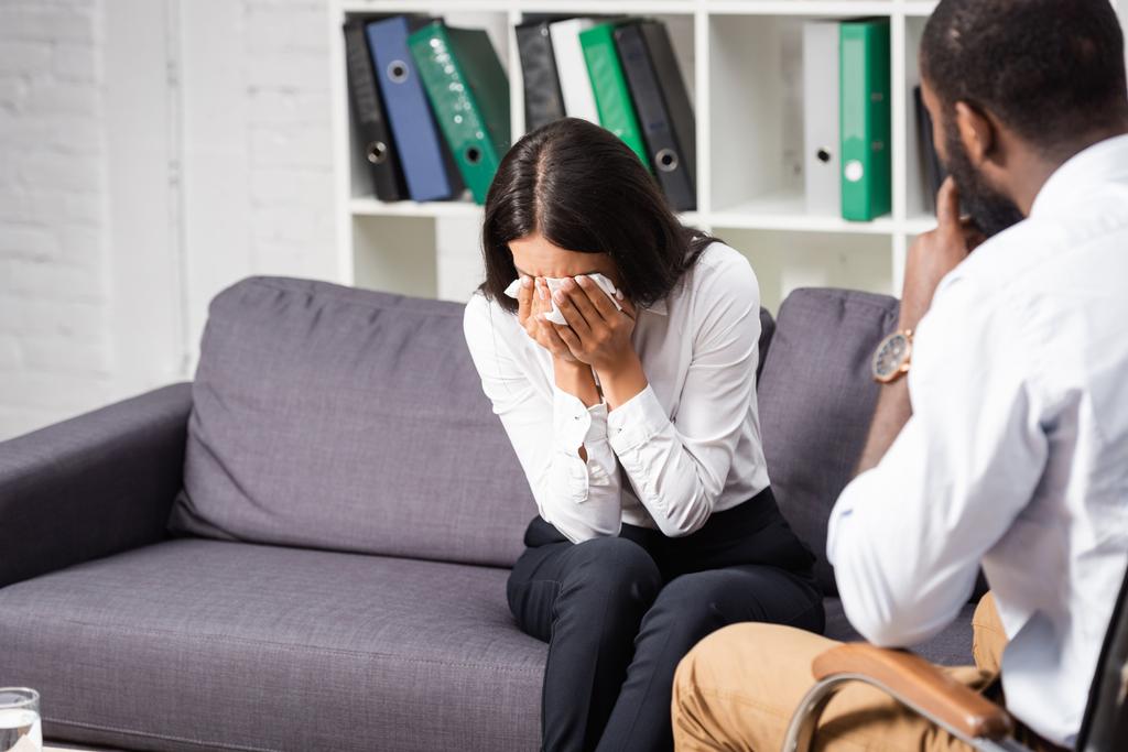 selectieve focus van Afrikaans-Amerikaanse psycholoog zit in de buurt van huilende vrouw vegen ogen met papieren servet - Foto, afbeelding