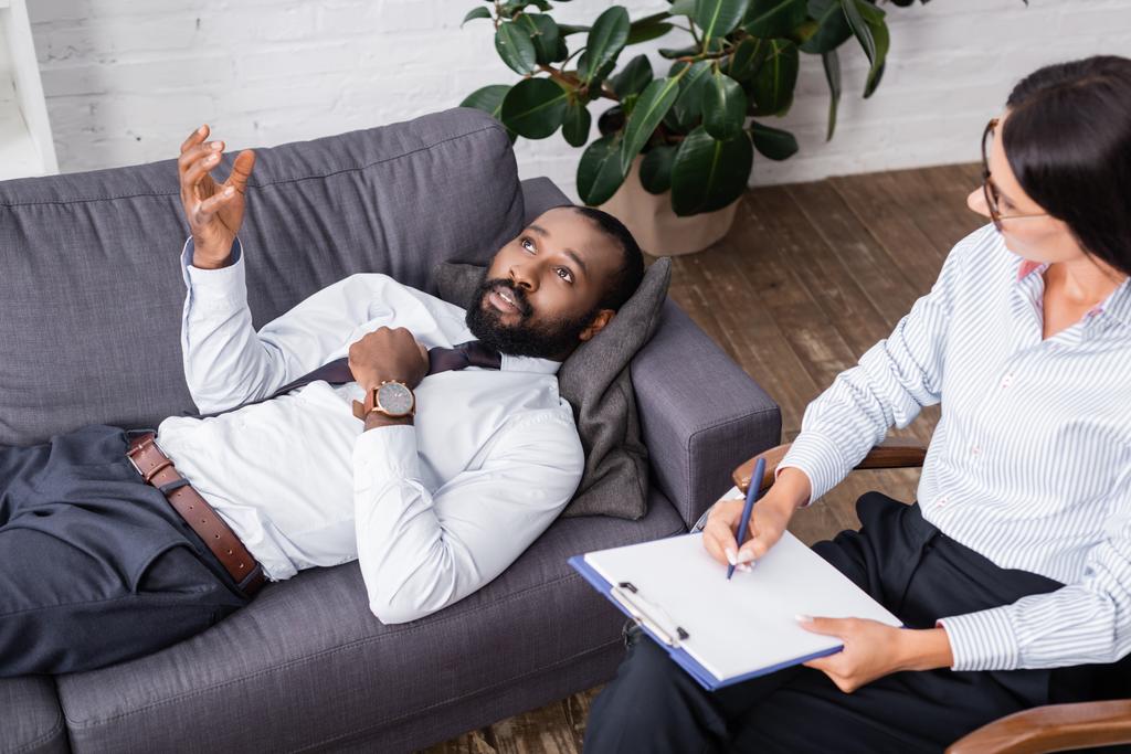 selektiver Fokus eines afrikanisch-amerikanischen Mannes, der gestikuliert und spricht, während er mit Stift und Klemmbrett auf dem Sofa neben einem Psychologen liegt - Foto, Bild