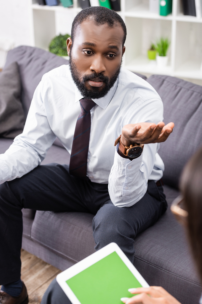 επιλεκτική εστίαση της τεταμένης Αφρικής Αμερικανός άνθρωπος gesturing, ενώ μιλάμε με ψυχολόγο κρατώντας ψηφιακή ταμπλέτα με λευκή οθόνη - Φωτογραφία, εικόνα