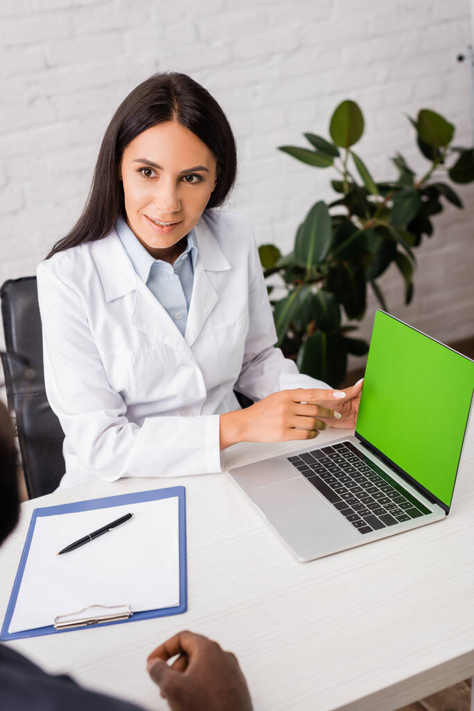 enfoque selectivo del médico apuntando con el dedo a la computadora portátil con pantalla verde cerca del paciente afroamericano  - Foto, Imagen