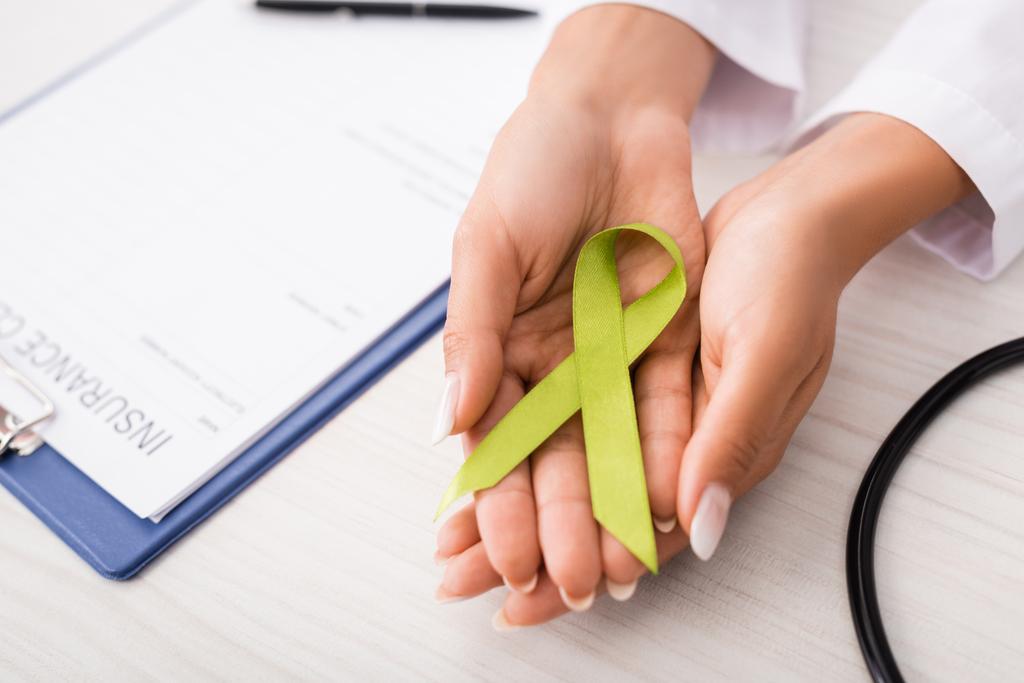 κορυφαία άποψη του γιατρού κρατώντας πράσινη κορδέλα ευαισθητοποίησης κοντά στο έντυπο αίτησης ασφάλισης, έννοια της ψυχικής υγείας - Φωτογραφία, εικόνα