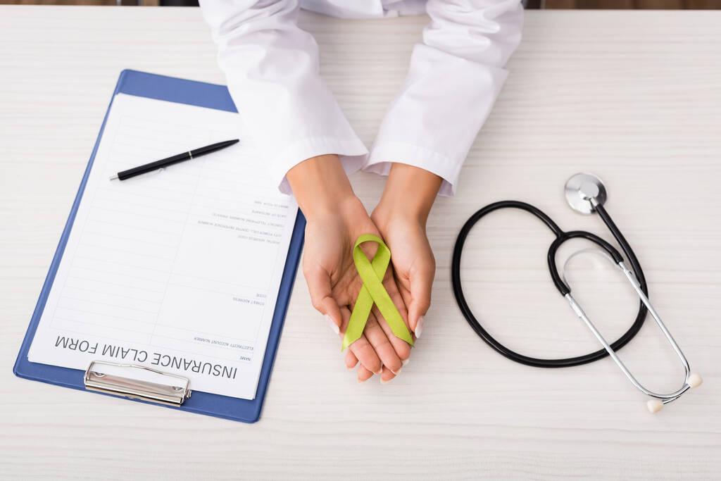 top view of psychologist holding green awareness ribbon near insurance claim form and stethoscope, mental health concept - Photo, Image
