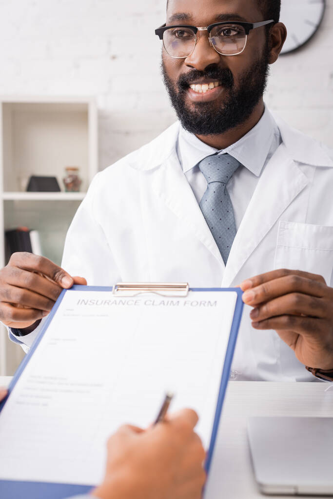 クリップボードを持っている楽しいアフリカ系アメリカ人医師の選択的焦点患者保険金請求書に署名しながら - 写真・画像