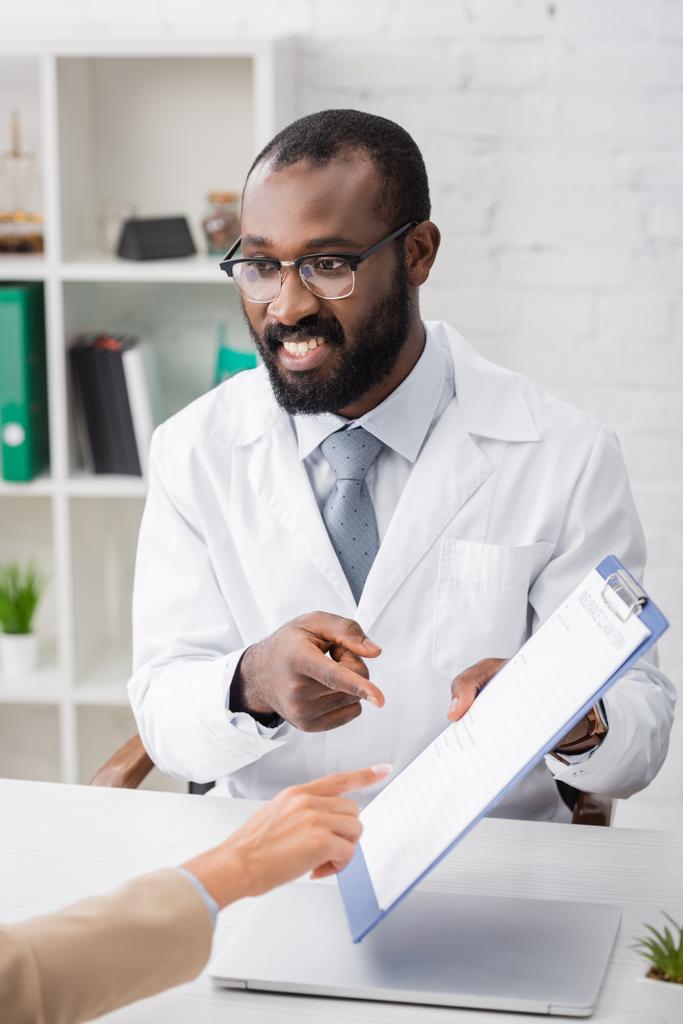 selektivní zaměření afroamerického lékaře a pacienta ukazujícího prsty na schránku s formulářem pojištění - Fotografie, Obrázek