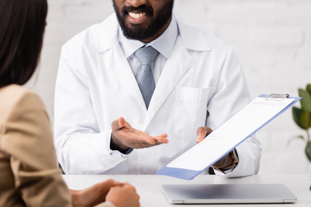 Ausgeschnittene Ansicht eines afrikanisch-amerikanischen Arztes, der mit dem Finger auf Klemmbrett mit Versicherungsformular in der Nähe des Patienten zeigt - Foto, Bild