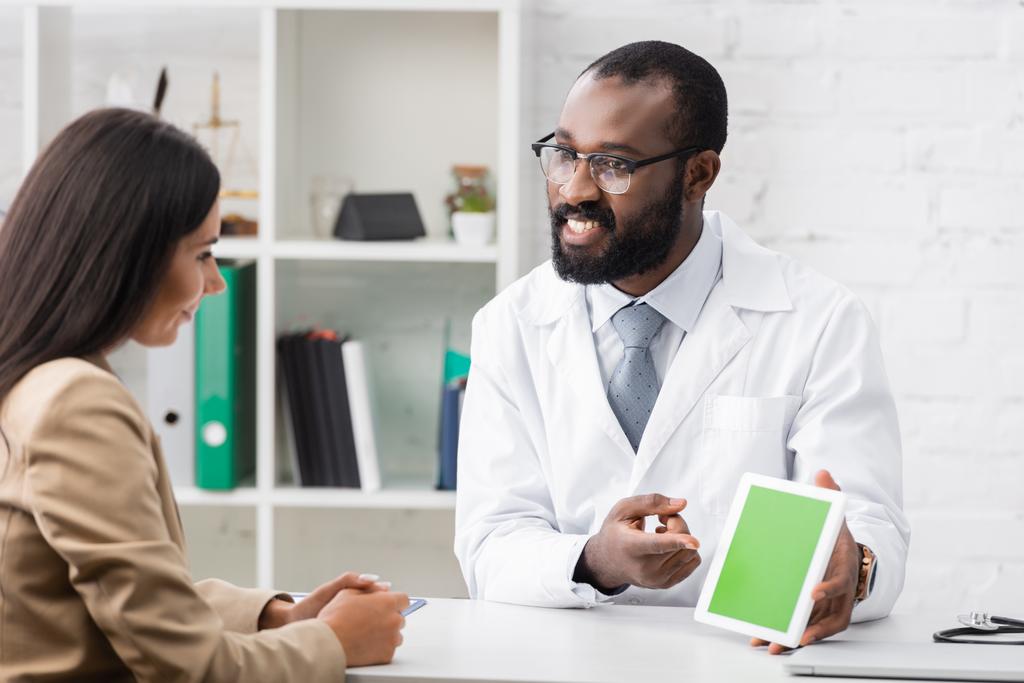vousatý africký americký lékař v brýlích ukazující prstem na digitální tabletu se zelenou obrazovkou poblíž brunetky pacient - Fotografie, Obrázek