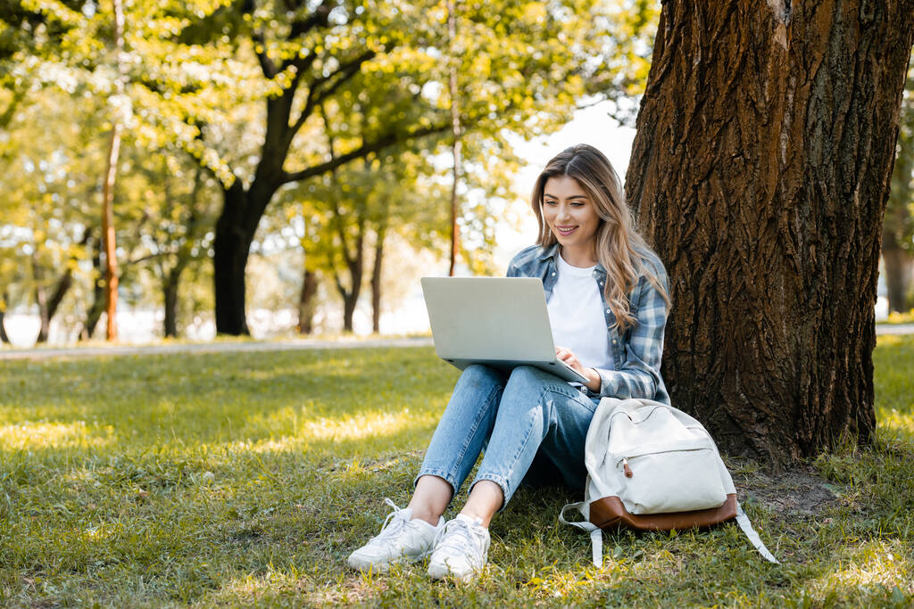 Kadın çimlerin üzerinde ağaç gövdesinin altında otururken dizüstü bilgisayar kullanıyor. - Fotoğraf, Görsel