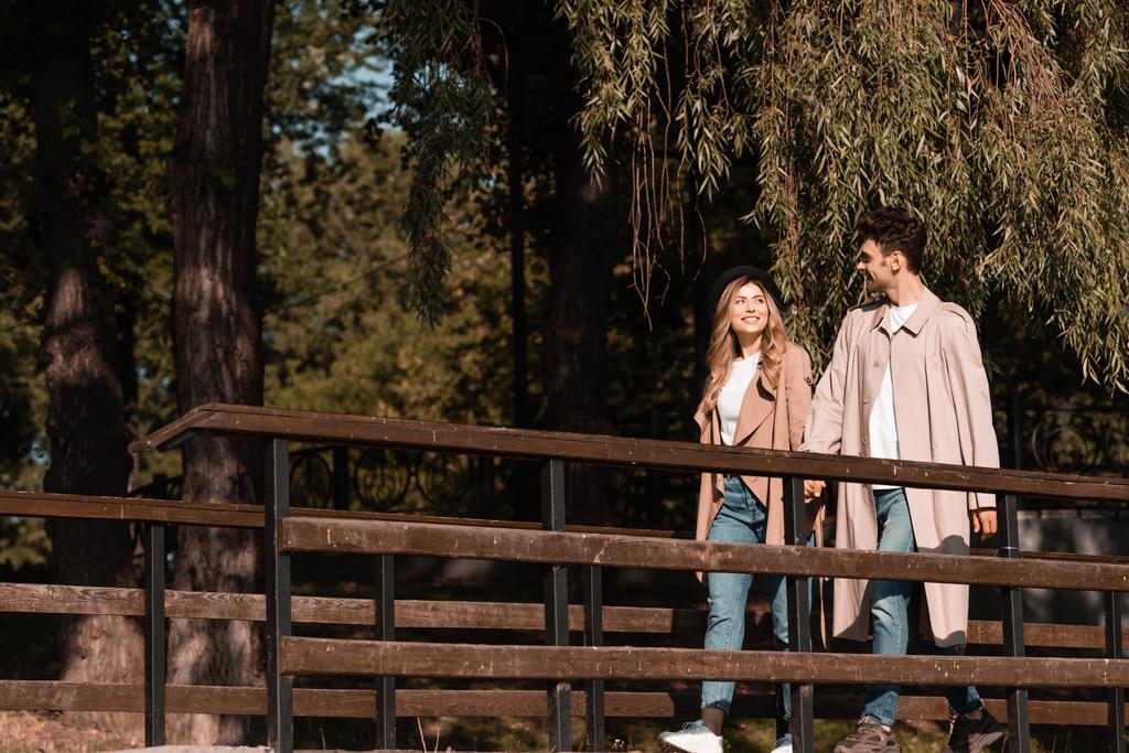 man and woman in trench coats holding hands and walking on wooden bridge - Photo, Image
