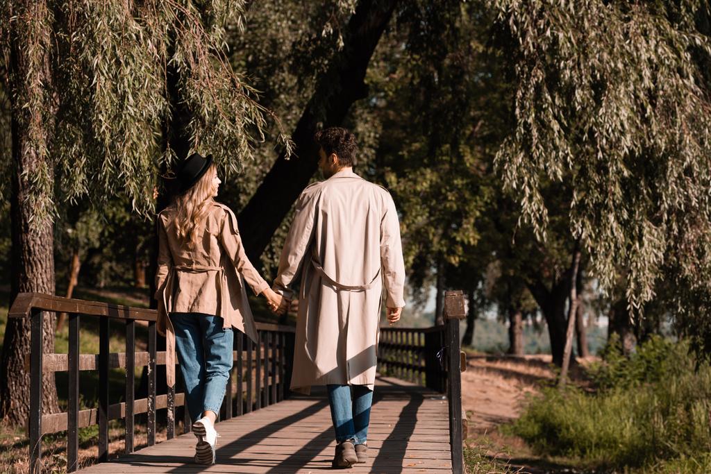 秋の公園で手を取り木製の橋を歩くトレンチコートのカップル - 写真・画像