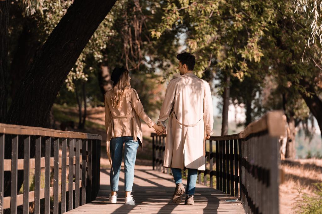 чоловік і жінка в траншеї пальто тримає руки, дивлячись один на одного і йдучи по дерев'яному мосту
 - Фото, зображення