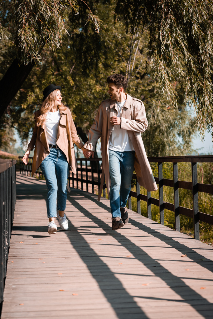 casal animado em casacos de trincheira de mãos dadas e andando na ponte de madeira - Foto, Imagem