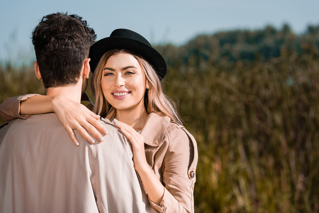 ξανθιά και χαρούμενη γυναίκα με μαύρο καπέλο που αγκαλιάζει άντρα με καμπαρντίνα  - Φωτογραφία, εικόνα